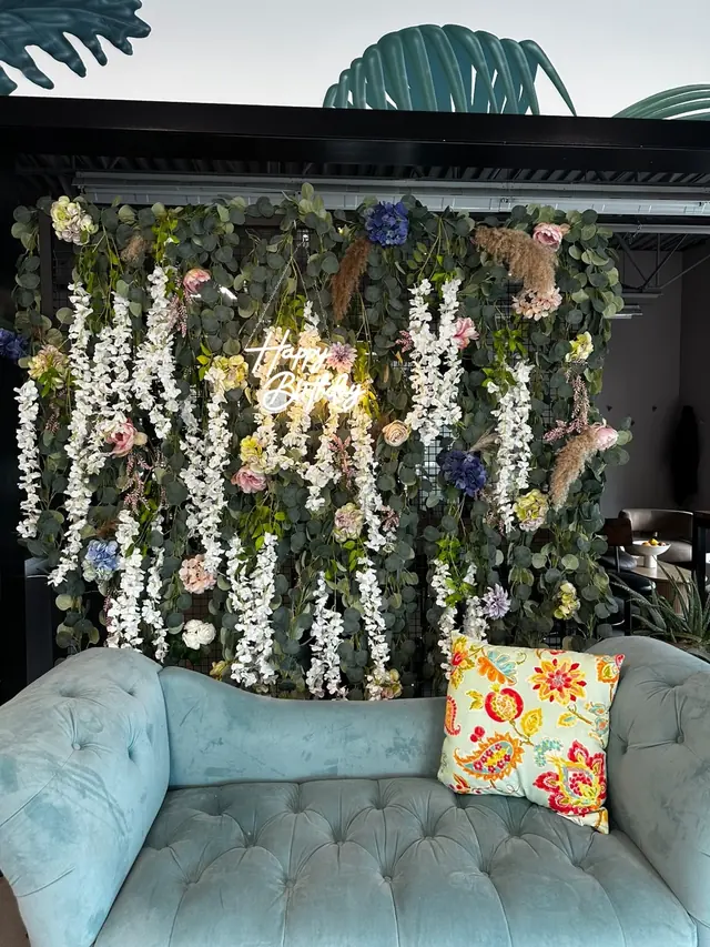 A hanging floral backdrop wall with a neon sign behind a teal velvet sofa